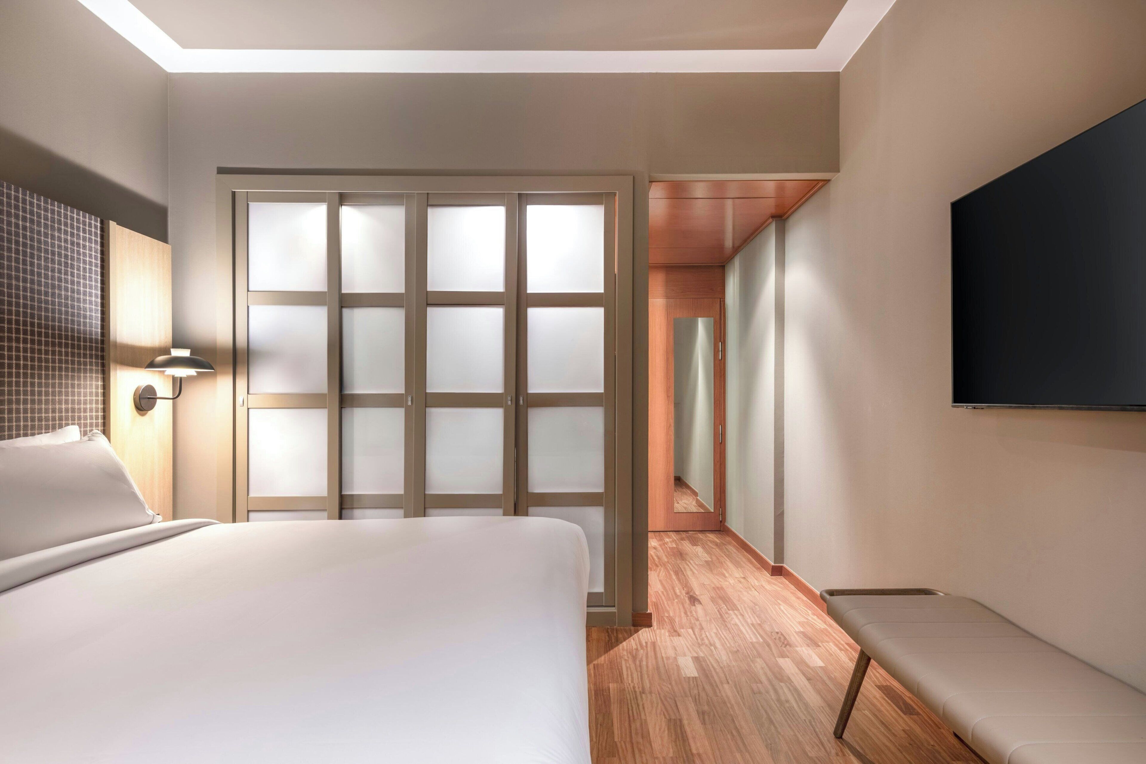 Quarto de hotel com cama, um banco, uma TV e uma porta de correr.