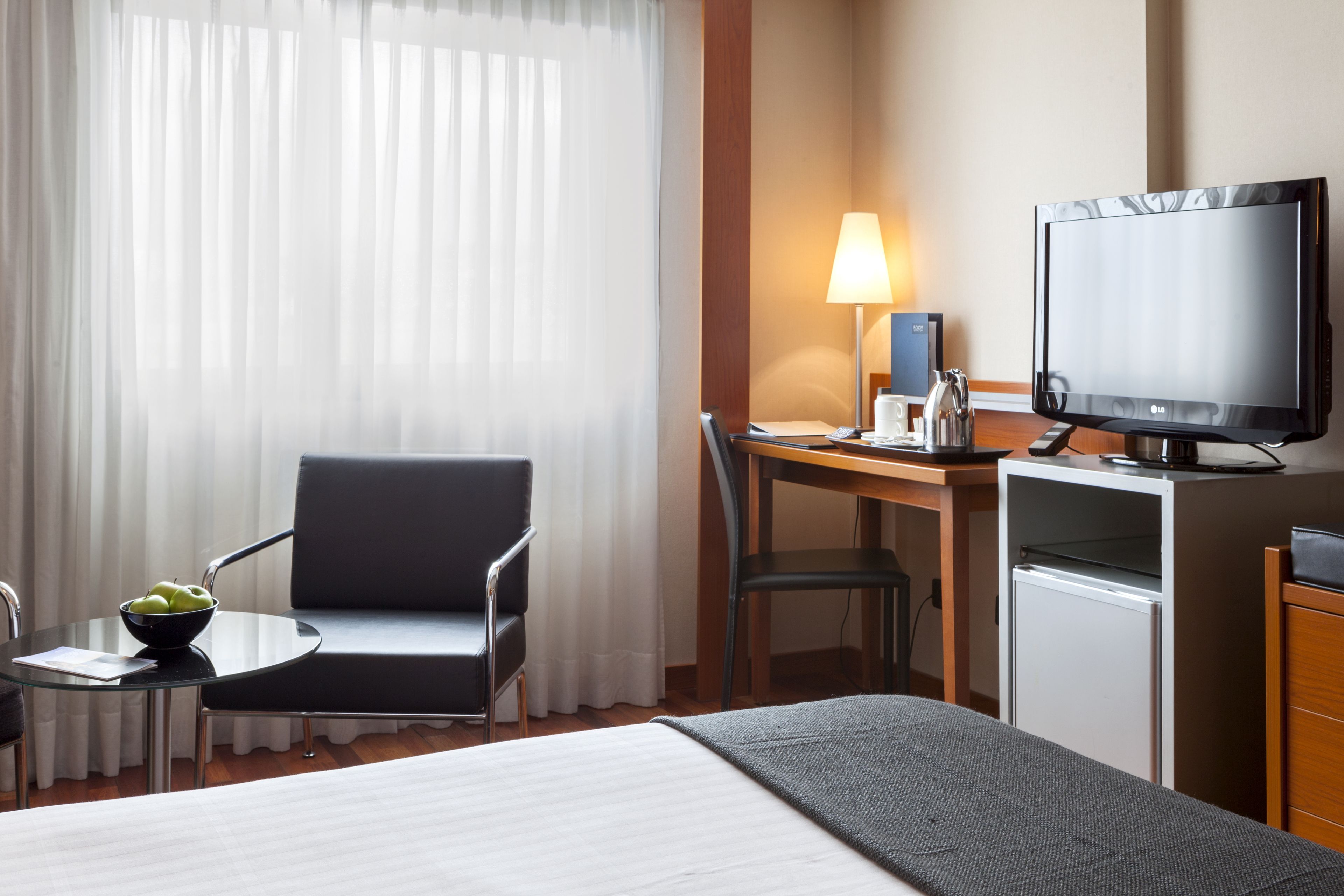 Ein Hotelzimmer mit einem Bett, einem Schreibtisch mit Computer und Lampe, einem Sessel, einem kleinen Tisch mit Obstschale und einem Fernseher.