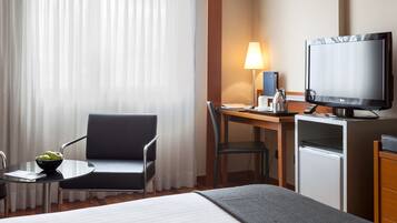 Ein Hotelzimmer mit einem Bett, einem Schreibtisch mit Computer und Lampe, einem Sessel, einem kleinen Tisch mit Obstschale und einem Fernseher.