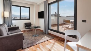 A modern living room with a sofa, a small table, and a chair. There is a large window with a view of buildings and a cityscape. - B&B Hotel Granada Estación (Granada)
