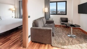 A hotel room with a bed, a sofa, a dining table, a TV, and a window with a city view. - B&B Hotel Granada Estación (Granada)