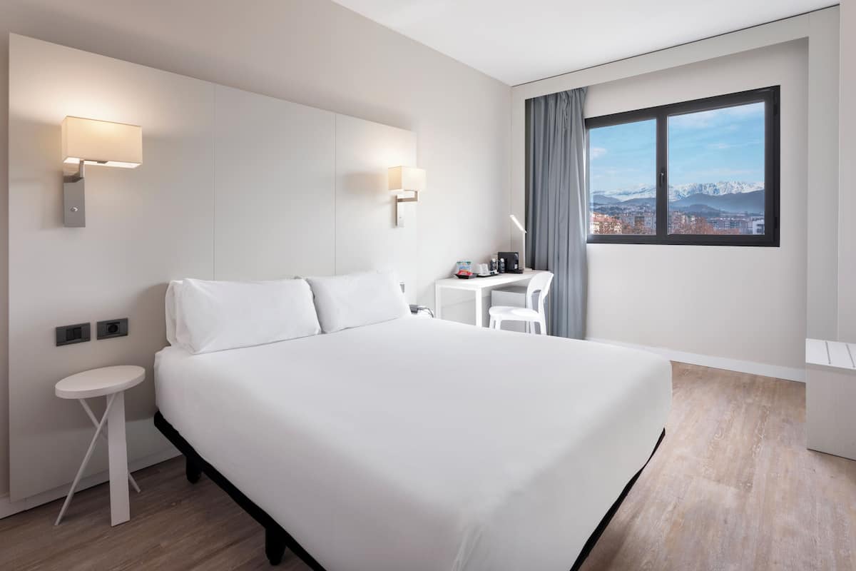 Et moderne hotelværelse med en stor seng, sengeborde, et skrivebord med stol og et vindue med udsigt over byen og bjergene.