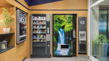 Zona de máquinas expendedoras con un mural de cascada, un dispensador de bebidas y una planta.