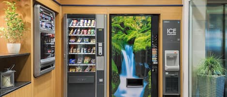 A vending machine area with a waterfall mural, a drink dispenser, and a plant.