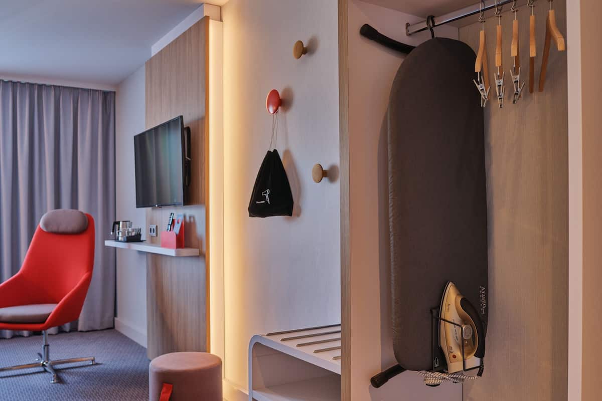 A modern hotel room with a red chair, a flat-screen TV, a wall-mounted ironing board, and a shelf with toiletries.