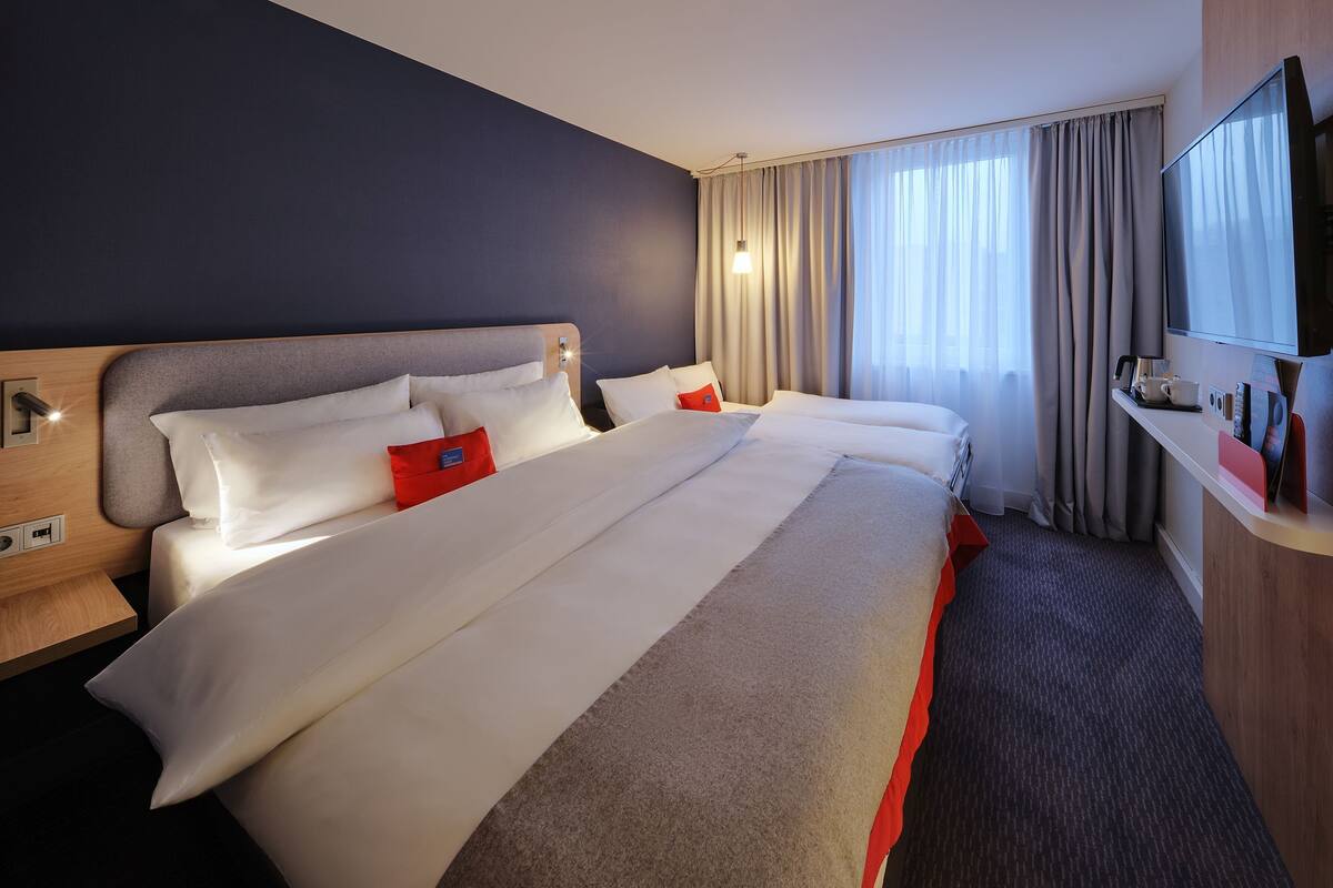 A hotel room with a large bed, a desk with a flat-screen TV, and a window with curtains.
