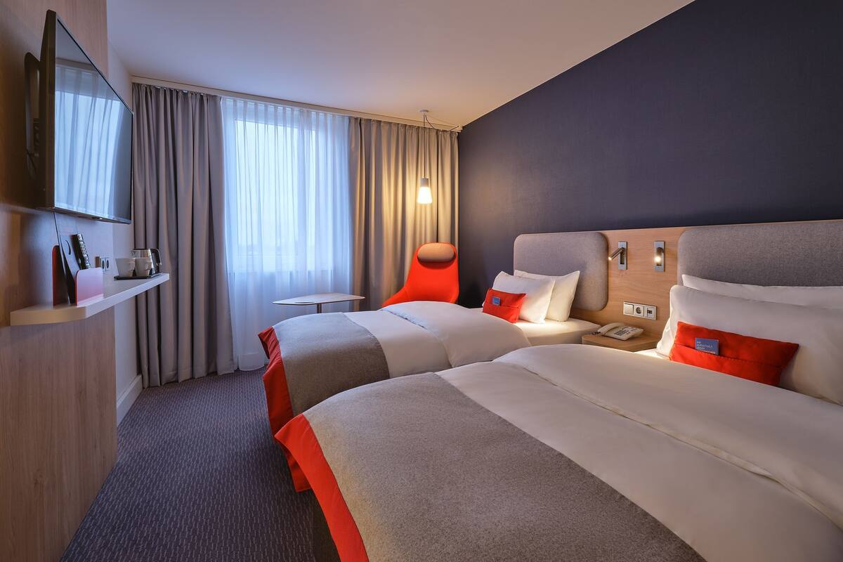 A hotel room with two beds, a TV, a desk, and a window with curtains.
