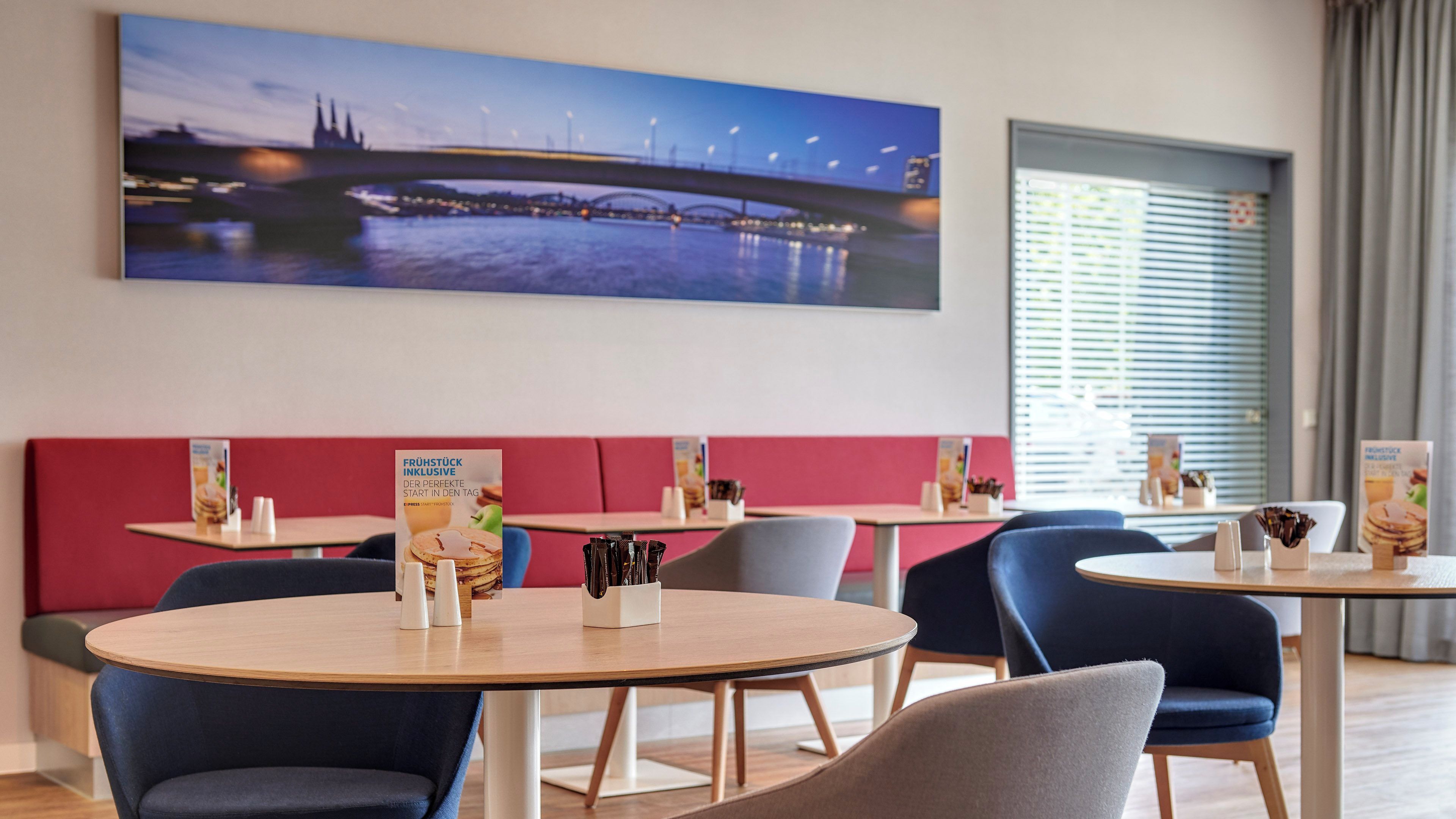 A modern dining area with round tables, red cushioned seating, and a cityscape painting on the wall.