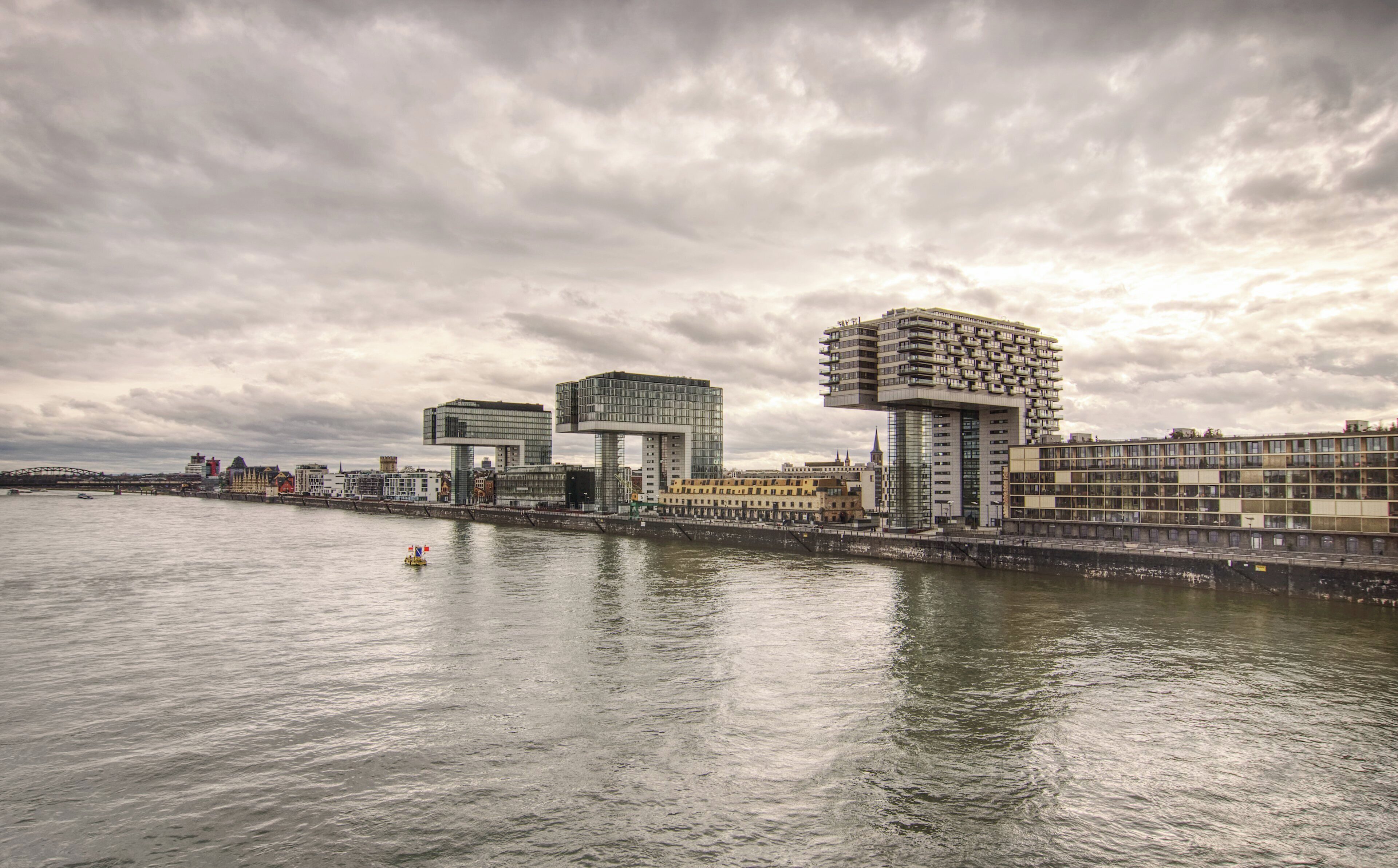 A waterfront cityscape with modern architecture, including a distinctive building with a square base and a rectangular top, and another with a grid-like facade.