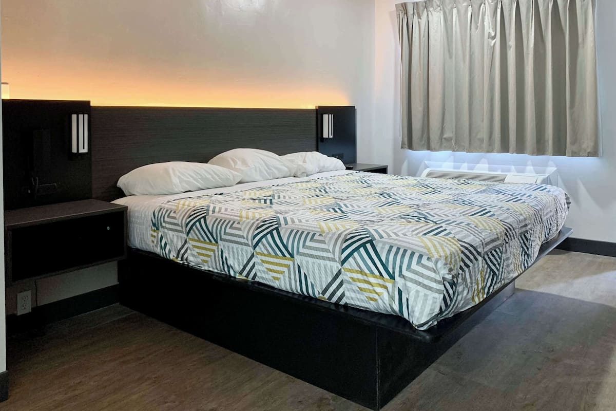 A modern hotel room with a large bed, dark headboard, bedside tables, and a window with curtains.