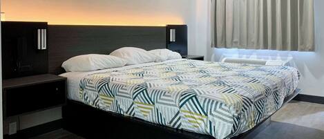 A modern hotel room with a large bed, dark headboard, bedside tables, and a window with curtains.