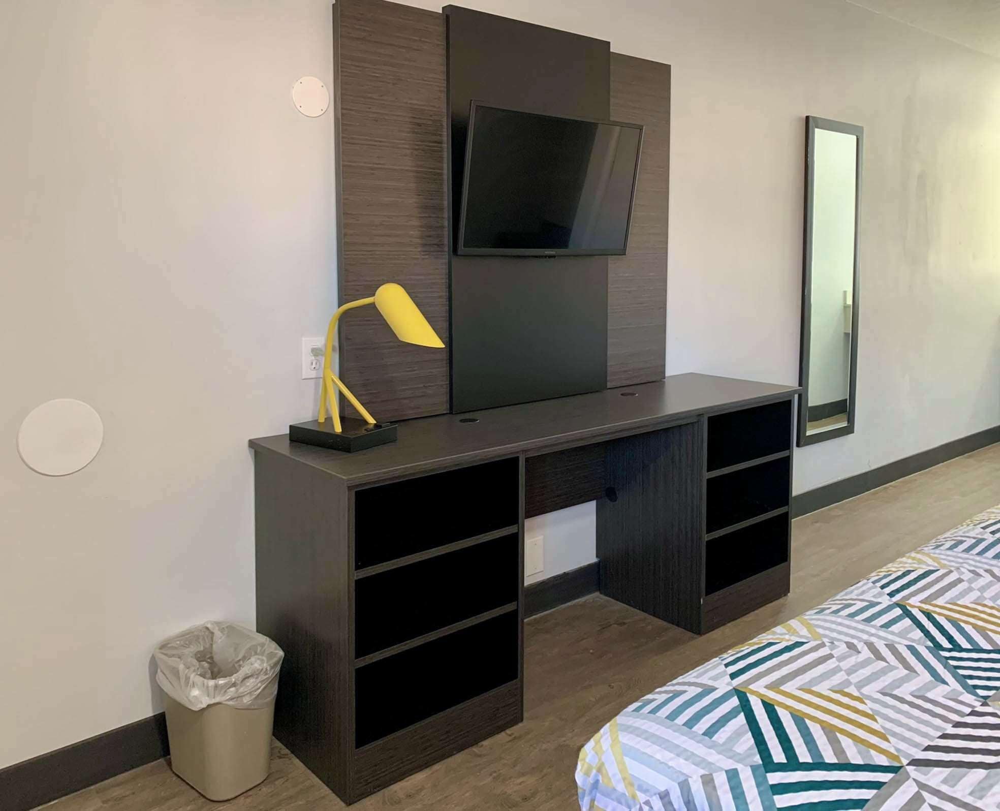 Habitación de hotel con un escritorio de madera oscura, un televisor de pantalla plana montado encima, una lámpara de escritorio amarilla pequeña, un cubo de basura y una cama con colcha estampada.