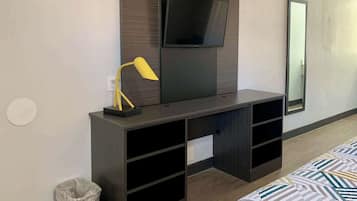 Habitación de hotel con un escritorio de madera oscura, un televisor de pantalla plana montado encima, una lámpara de escritorio amarilla pequeña, un cubo de basura y una cama con colcha estampada.