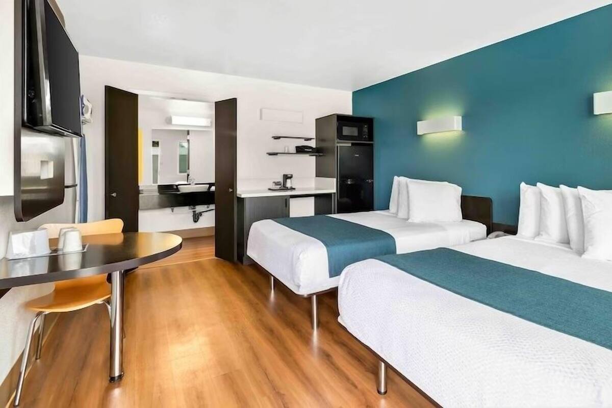 A hotel room with two beds, a desk, a chair, a TV, a microwave, and a bathroom.