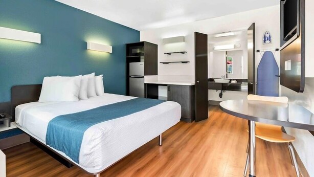 Habitación de hotel con cama, escritorio, silla, espejo y kitchenette.