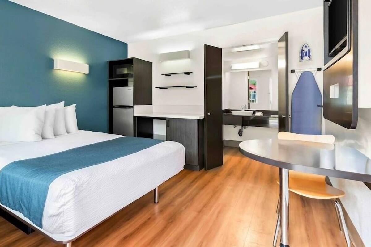 A hotel room with a bed, a desk, a chair, a mirror, and a kitchenette.