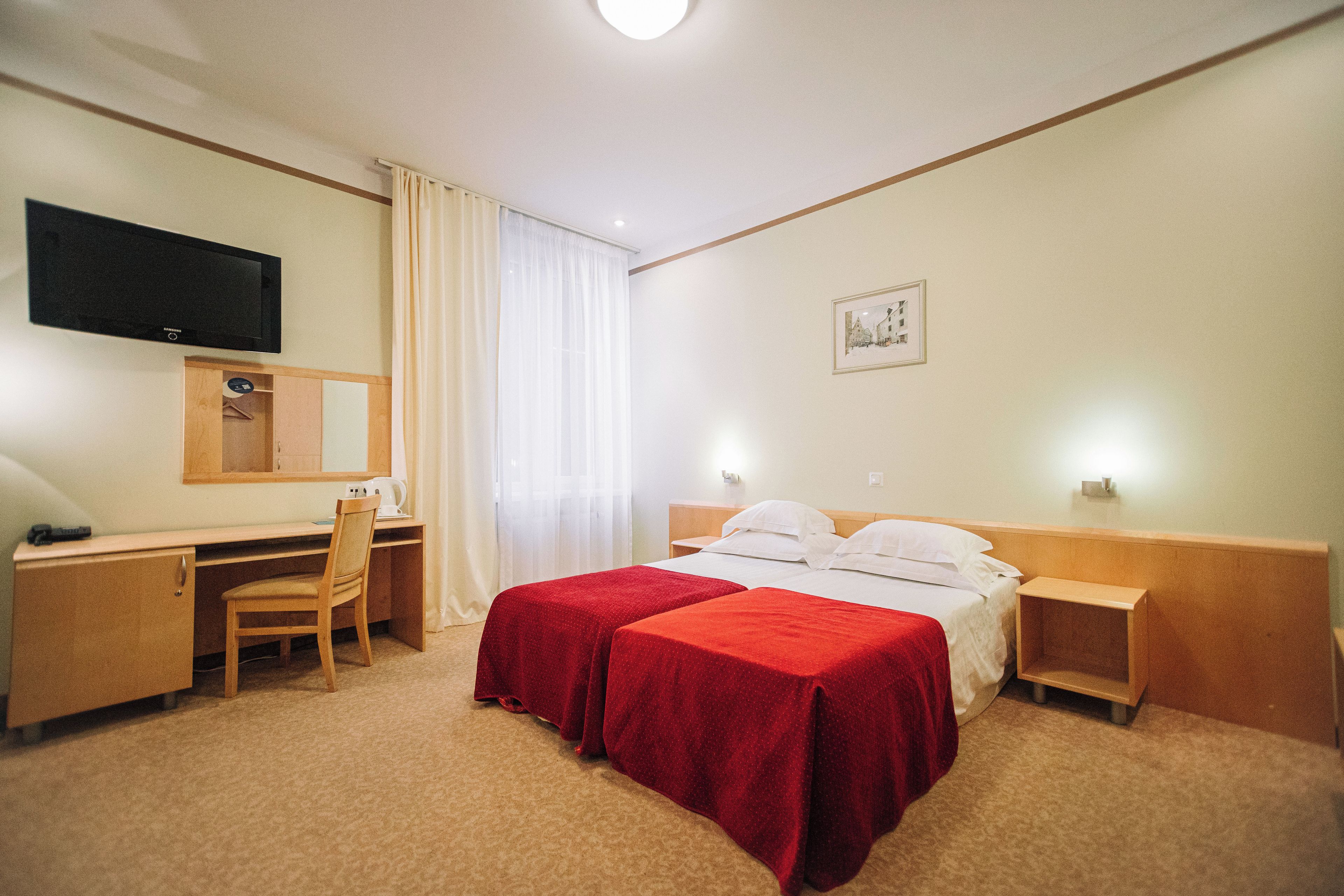 A hotel room with a bed, two bedside tables, a desk with a chair, a television mounted on the wall, and a window with curtains.