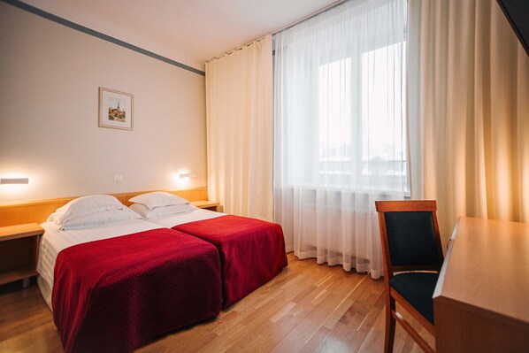 Habitación de hotel con dos camas individuales, una mesita de noche de madera, un escritorio, una silla y un ventanal con cortinas translúcidas.
