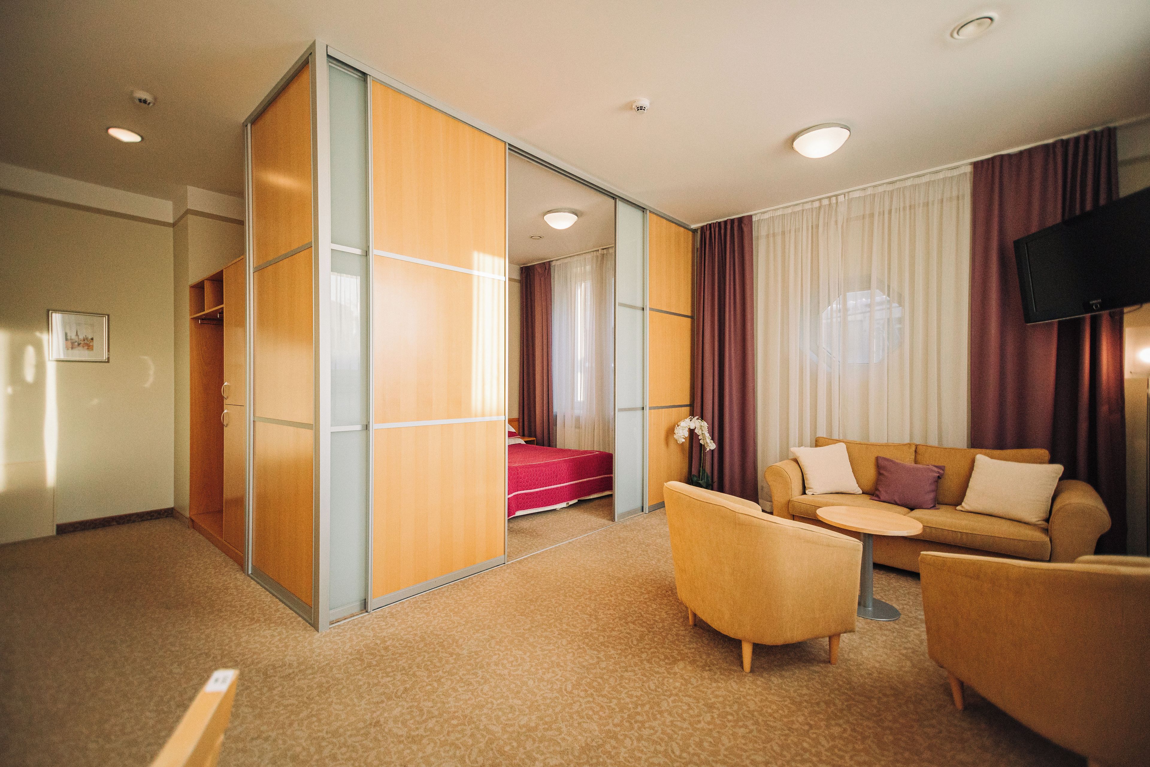 A hotel room with a sofa, armchairs, a TV, a sliding door, and a bed with a red cover.