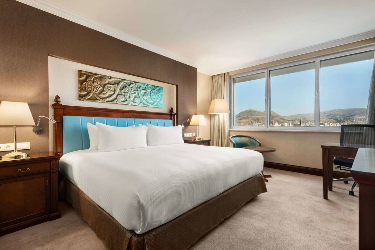 A hotel room with a large bed, two bedside tables, a desk, and a chair. There is a large window with a view of a cityscape.