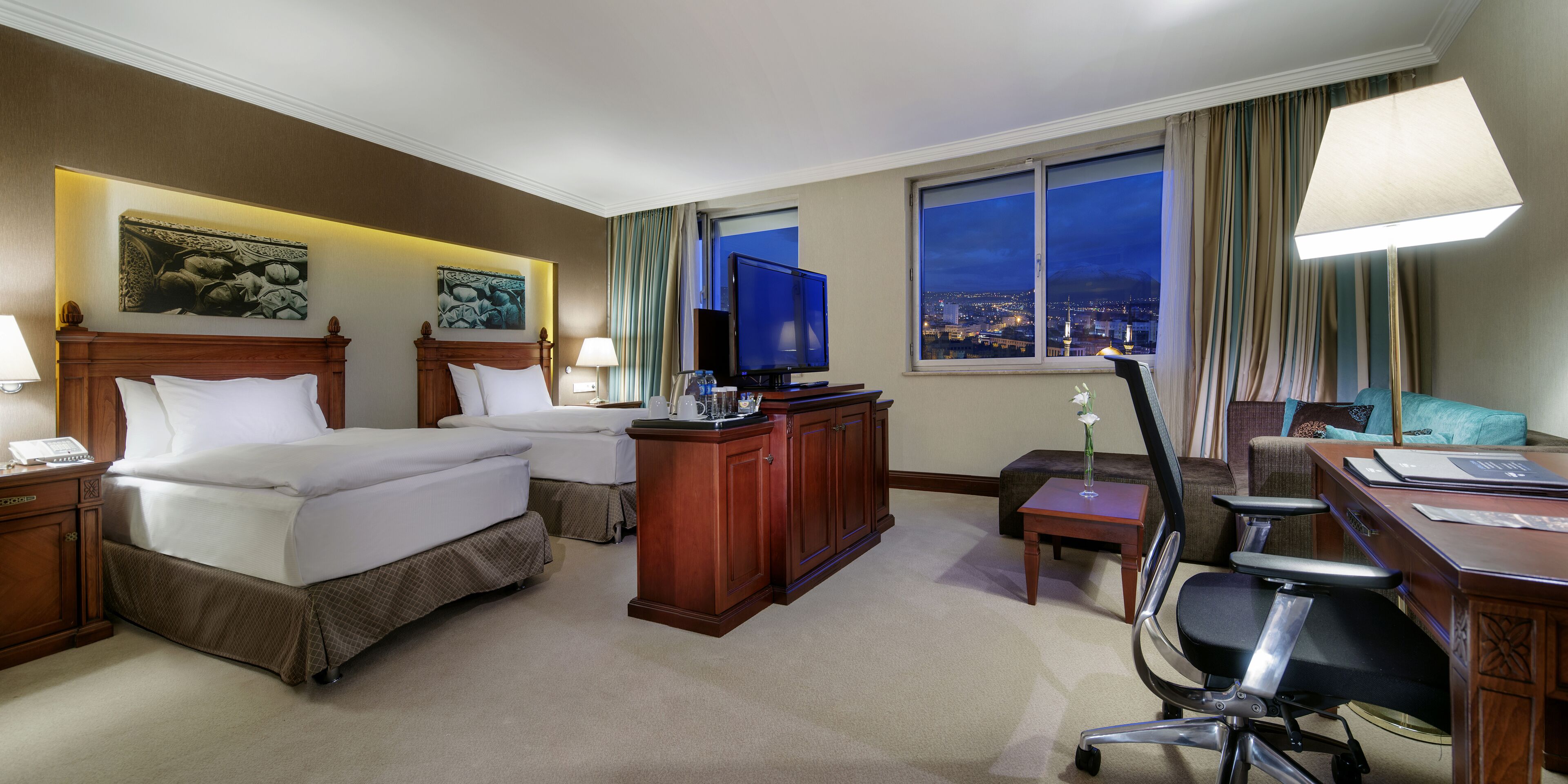 A hotel room with two beds, a desk, a chair, a television, and a view of the city at night.
