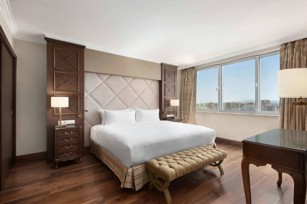 A hotel room with a large bed, a wooden nightstand, a bench, a desk, and a view of the city through large windows.