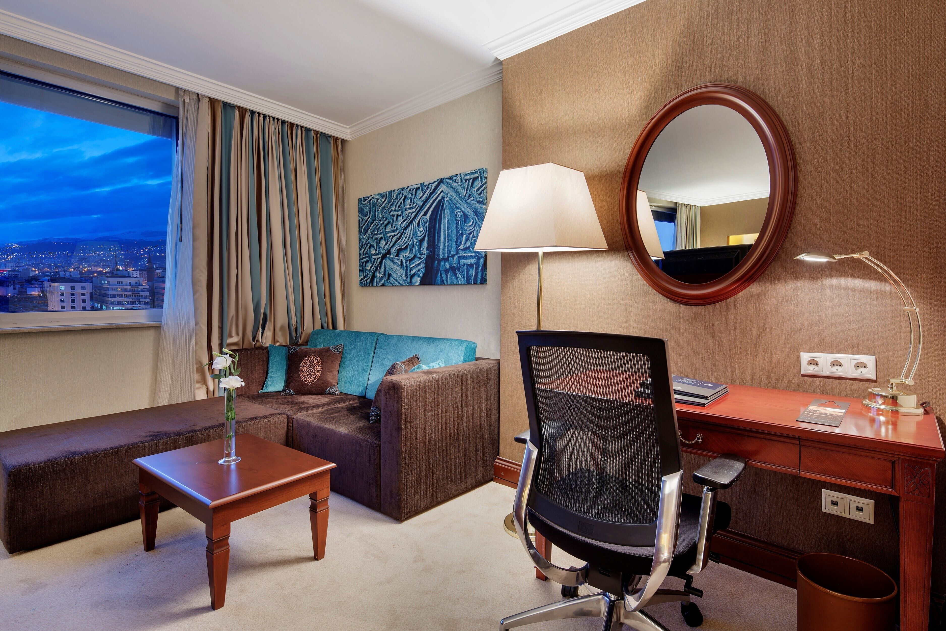 A hotel room with a sofa, a desk, a chair, a lamp, a mirror, and a window with a city view.