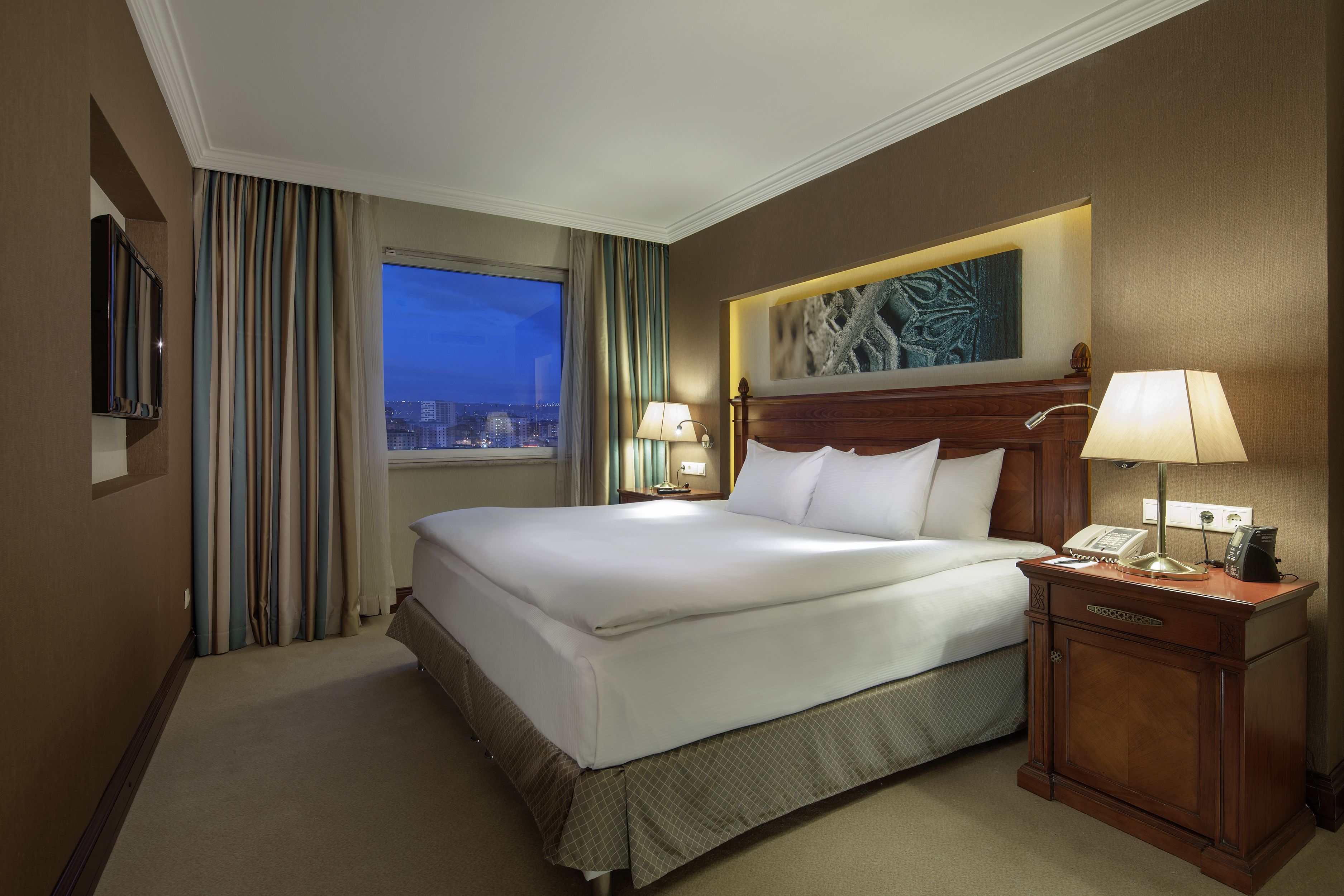 A hotel room with a large bed, two bedside tables with lamps, a flat-screen TV, and a window with curtains.