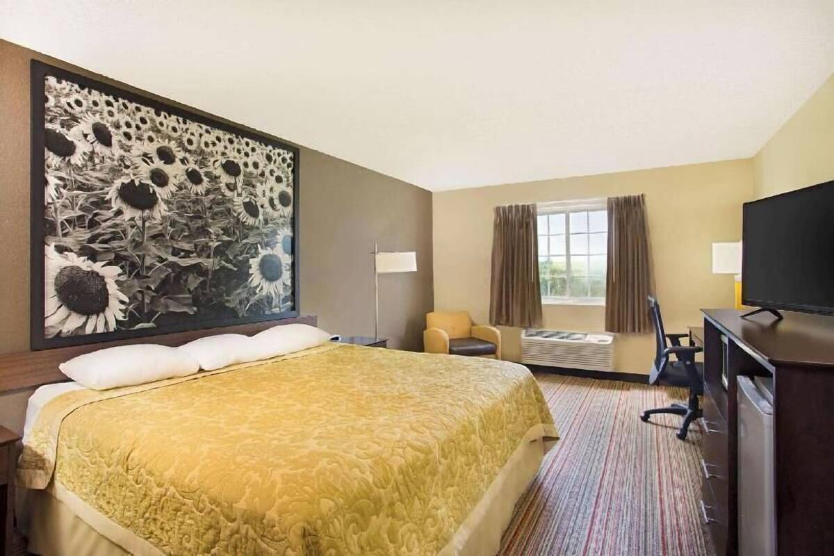 Ein Hotelzimmer mit einem großen Bett, einem Schreibtisch, einem Stuhl, einem Fernseher und einem großen Gemälde von Sonnenblumen an der Wand.