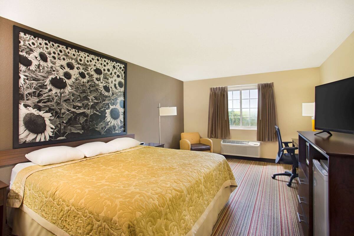 Ein Hotelzimmer mit einem großen Bett, einem Schreibtisch mit Stuhl, einem Fernseher und einem Gemälde von Sonnenblumen an der Wand.