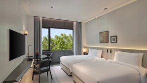 Habitación de hotel con dos camas, televisión, escritorio con silla y ventana con vista a árboles.