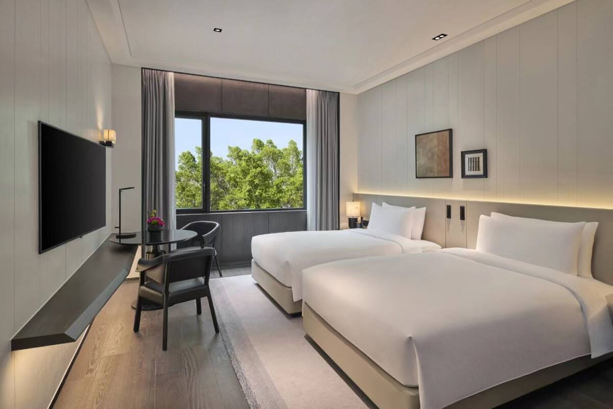 Une chambre d’hôtel avec deux lits, une télévision, un bureau avec une chaise et une fenêtre donnant sur des arbres.