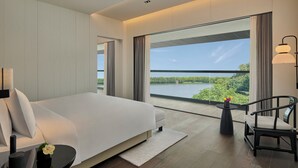 Una habitación de hotel moderna con una cama grande, un área de descanso con una silla, una mesita y vistas a un lago a través de amplios ventanales.
