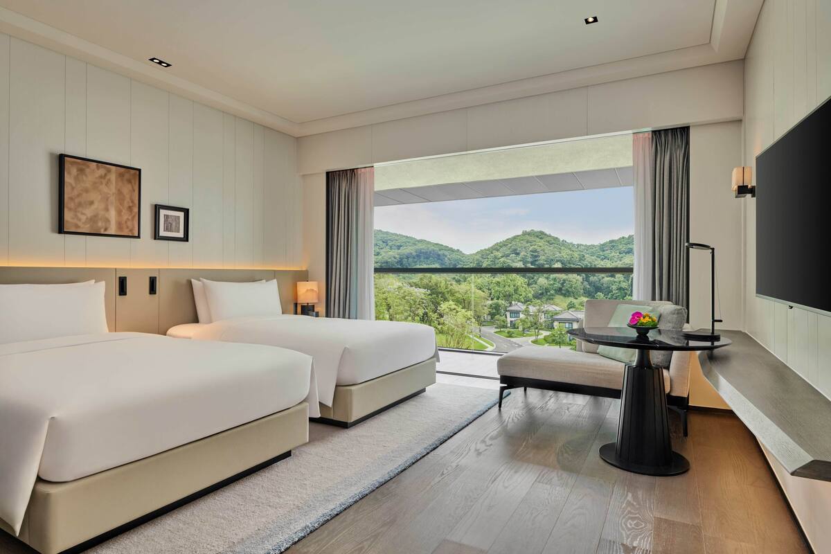 Une chambre d’hôtel avec deux lits, un bureau, une télévision et une grande fenêtre donnant sur un paysage verdoyant.