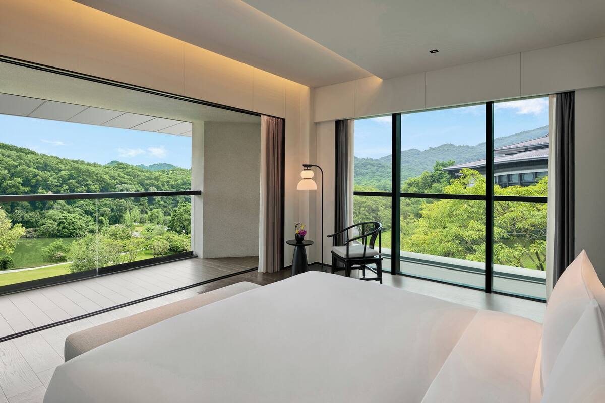 Une chambre d’hôtel moderne, dotée d’un grand lit, d’une chaise, d’une petite table et offrant une vue sur un paysage verdoyant grâce à de grandes fenêtres.