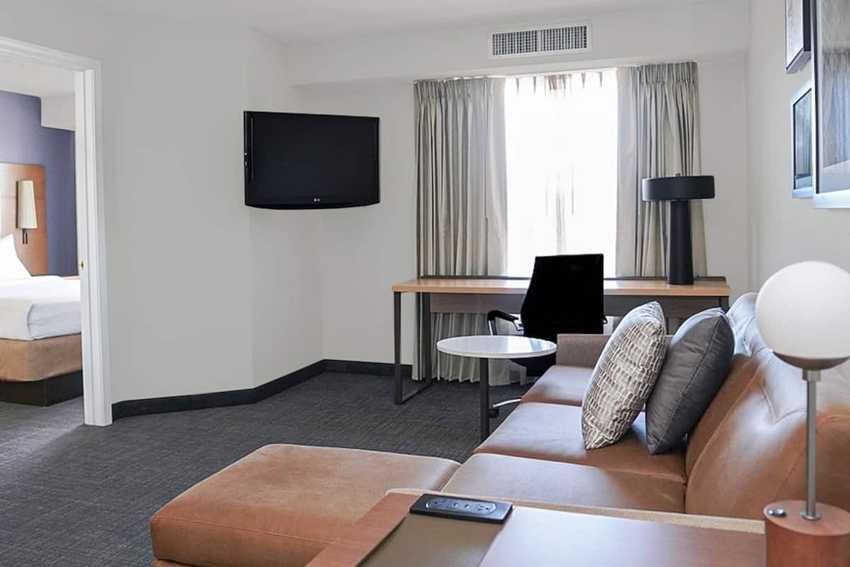 A hotel room with a brown leather sofa, a small round table, a desk with a chair, a flat-screen TV mounted on the wall, and a window with curtains.