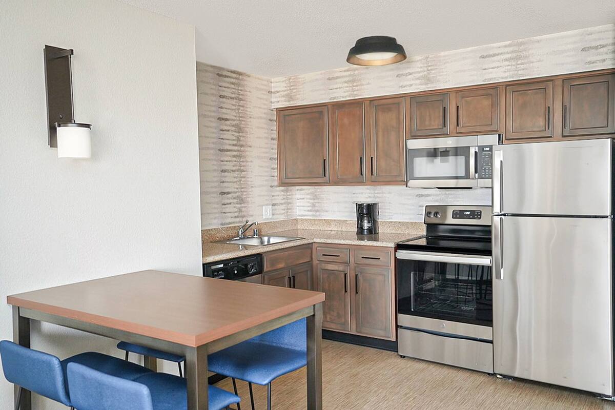 A compact kitchen with wooden cabinets, a stainless steel refrigerator, a microwave, and an oven. There is a small dining table with blue chairs.
