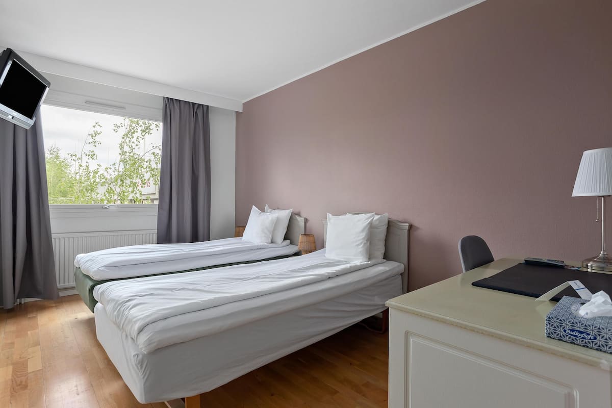 Ein Zimmer mit zwei Betten, einem Schreibtisch, einer Lampe und einem Fenster mit Vorhängen.