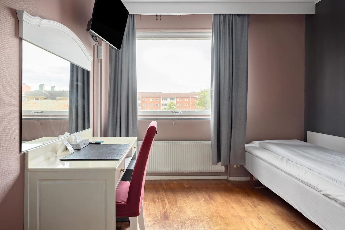 Ein Schlafzimmer mit Bett, Schreibtisch mit Stuhl, Fenster mit Vorhängen und Heizkörper.