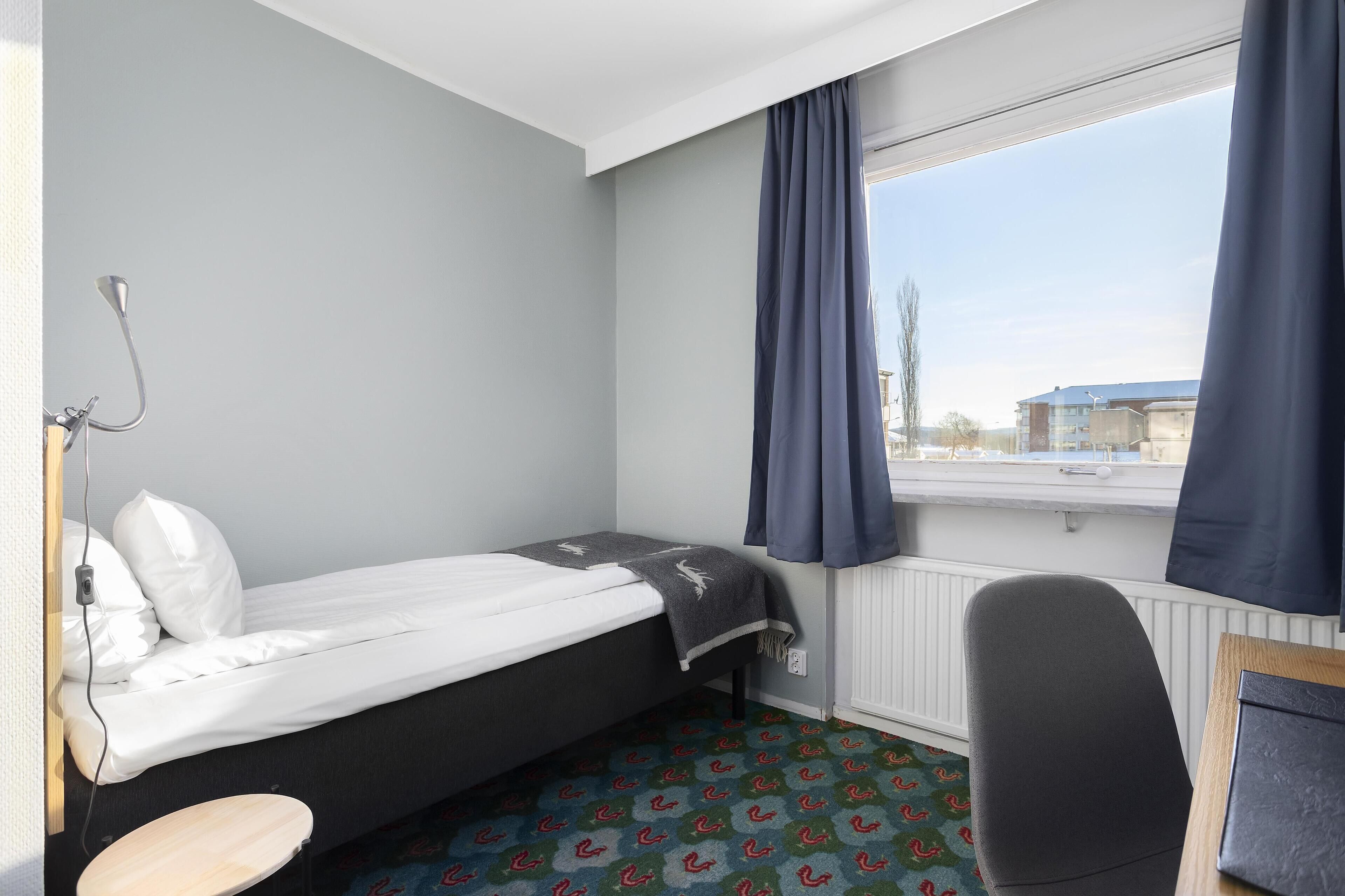 Una habitación de hotel pequeña con una cama individual, un escritorio y una ventana con cortinas.
