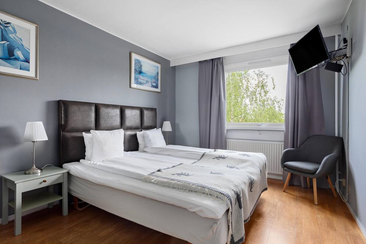Ein Hotelzimmer mit einem Bett, einem Nachttisch, einem Sessel, einem an der Wand montierten Fernseher und zwei gerahmten Bildern an der Wand.