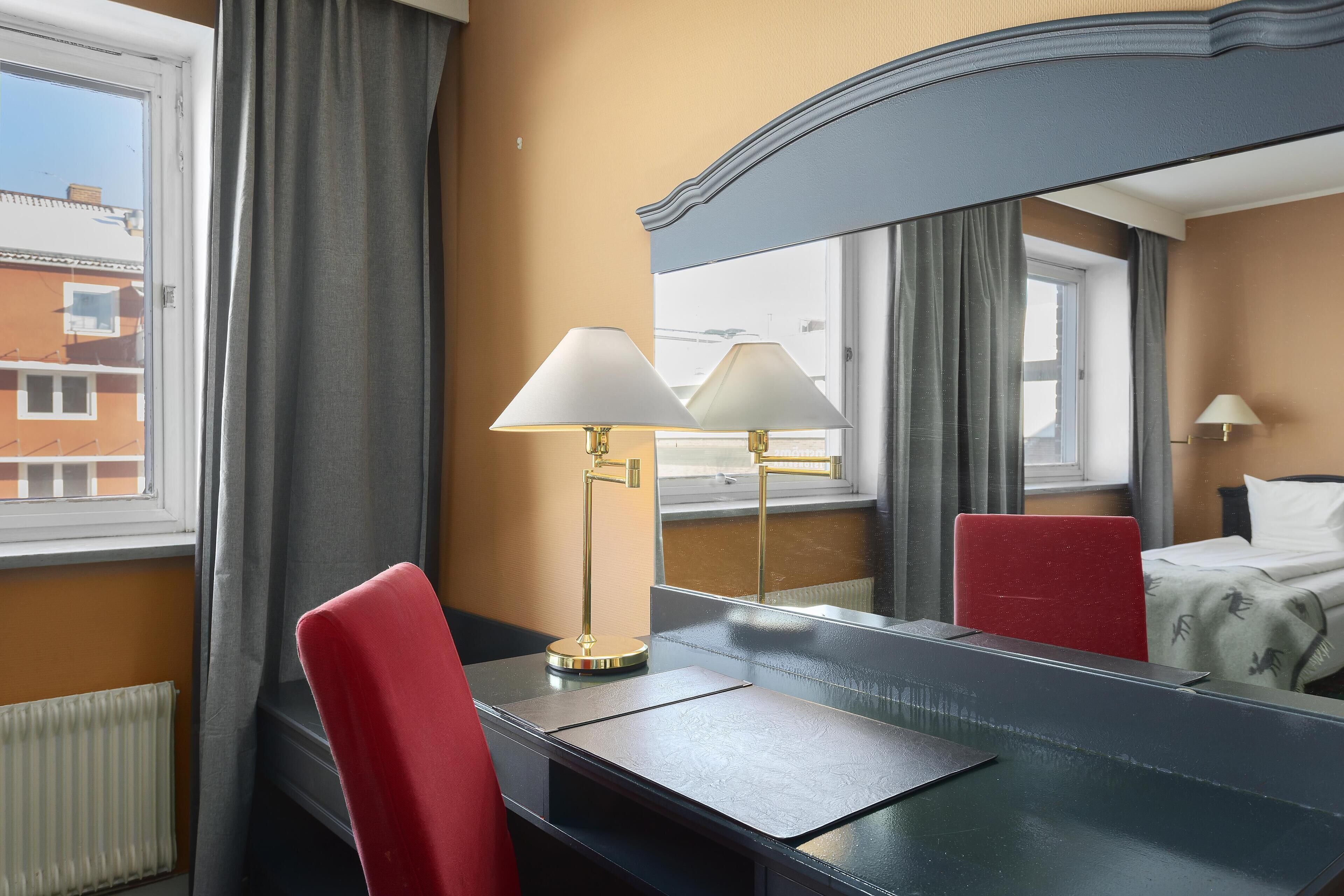 Ett hotellrum med ett skrivbord, två lampor, en spegel, en säng och ett fönster med gardiner.