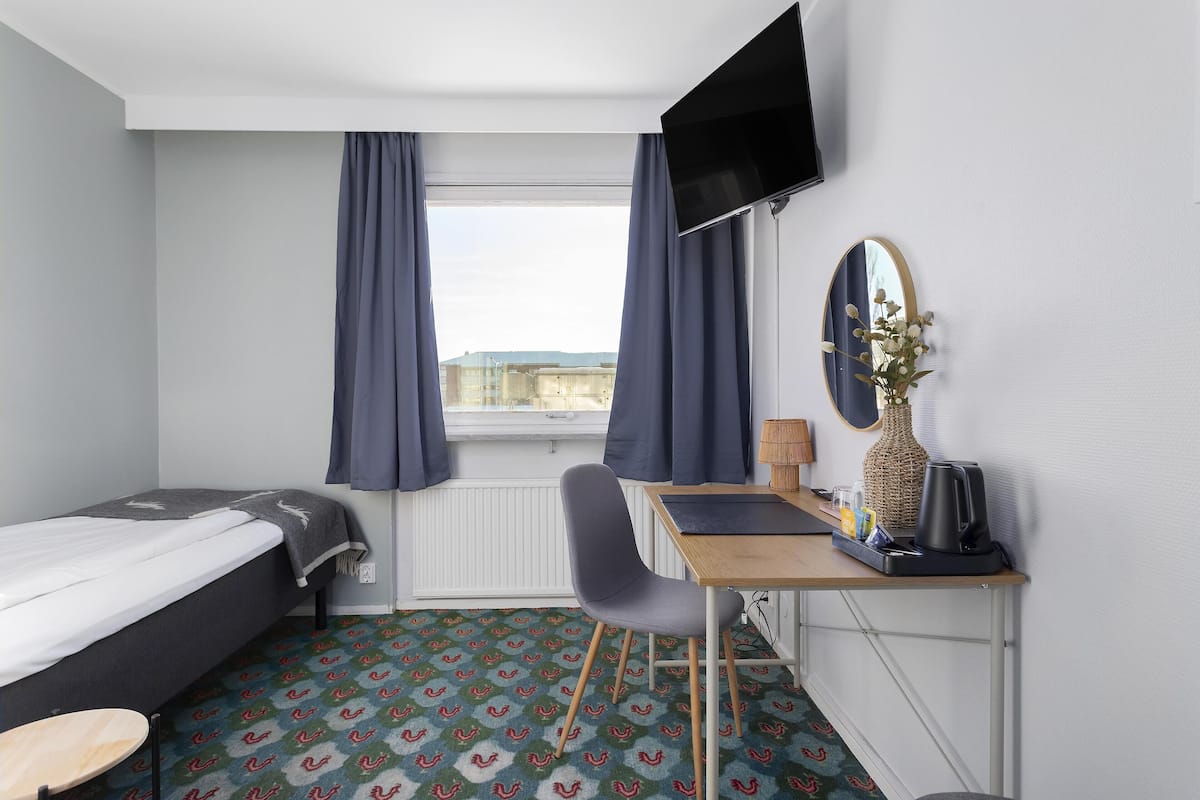 Ein Hotelzimmer mit einem Bett, einem Schreibtisch mit Stuhl, einem Fernseher, einem Spiegel und einem Fenster mit Vorhängen.