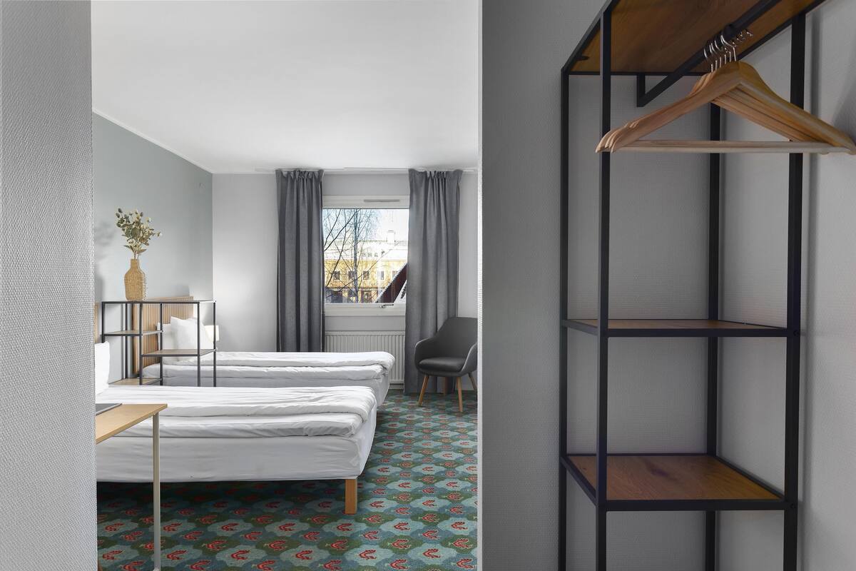 Ein Hotelzimmer mit zwei Betten, einem Sessel, einem hölzernen Kleiderbügel und einem Fenster mit Vorhängen.