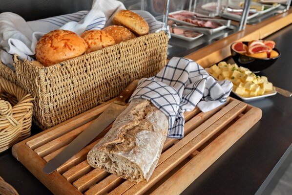 A wooden cutting board with a loaf of bread, a basket of bread rolls, and a bowl of cheese cubes. - Best Western Hotel Bollnas (Bollnas)