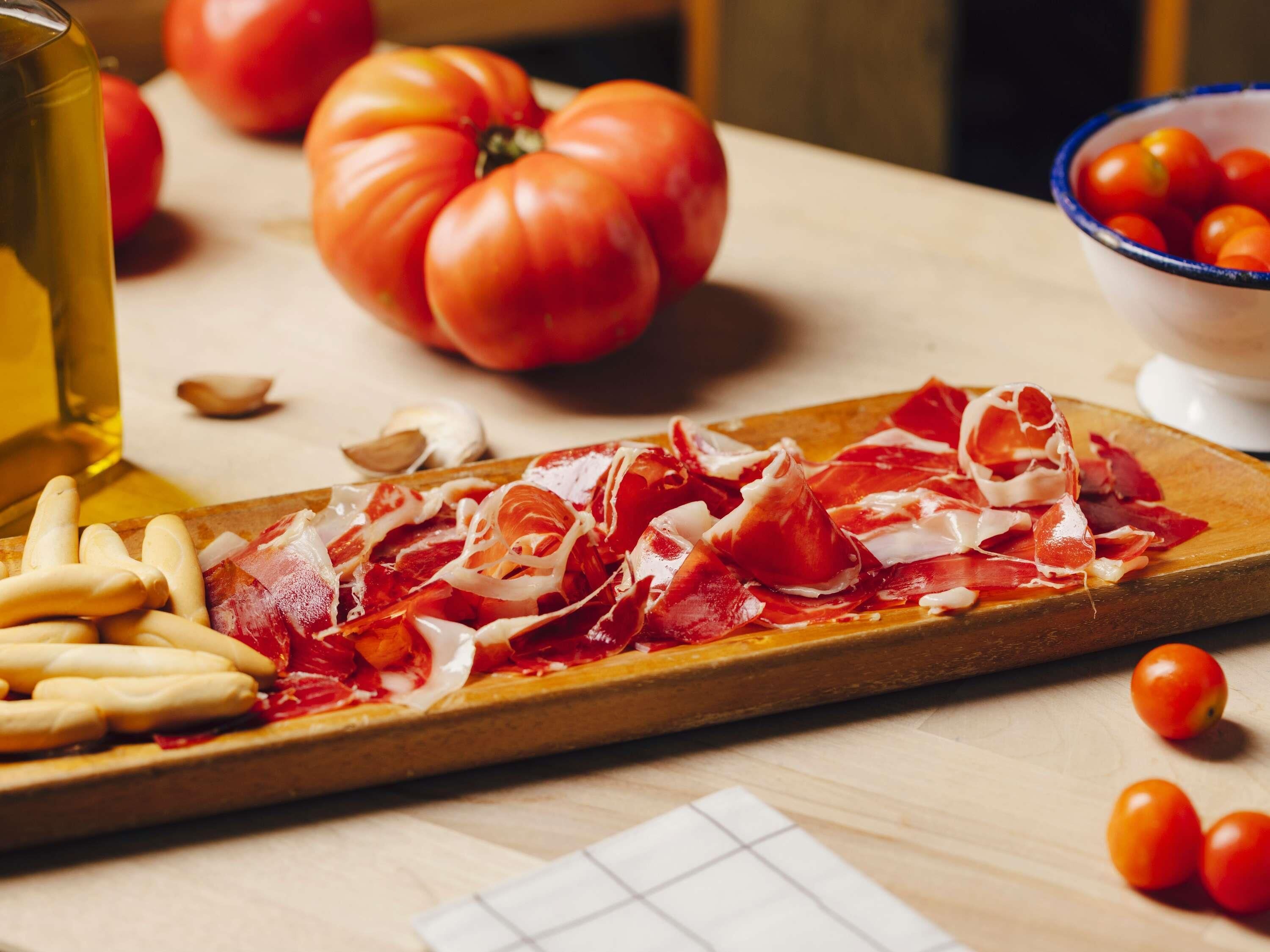 Una tabla de madera con carne curada en rodajas y un tazón de tomates cherry.