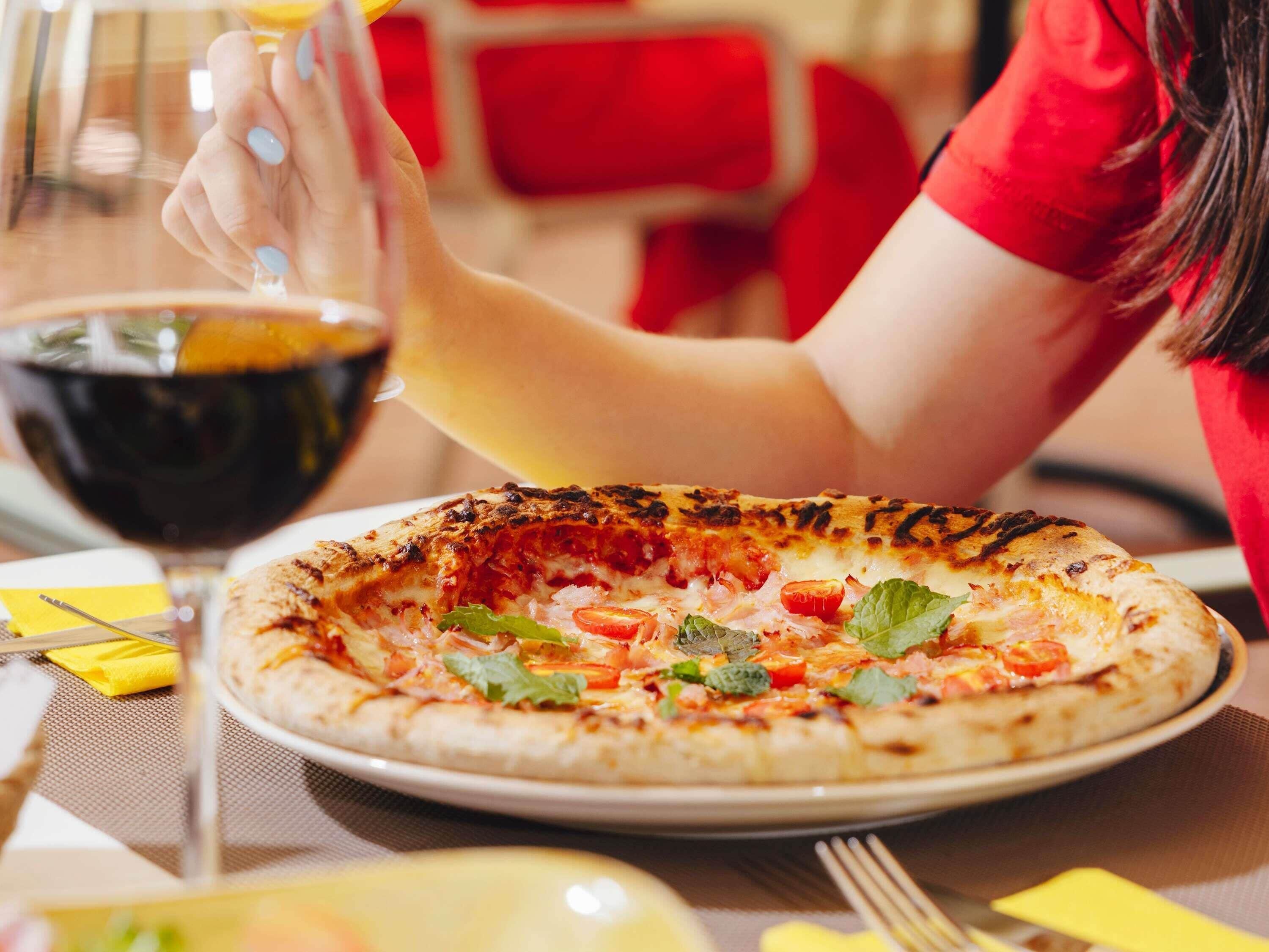 Une pizza garnie, un verre de vin rouge et une personne portant un t-shirt rouge.