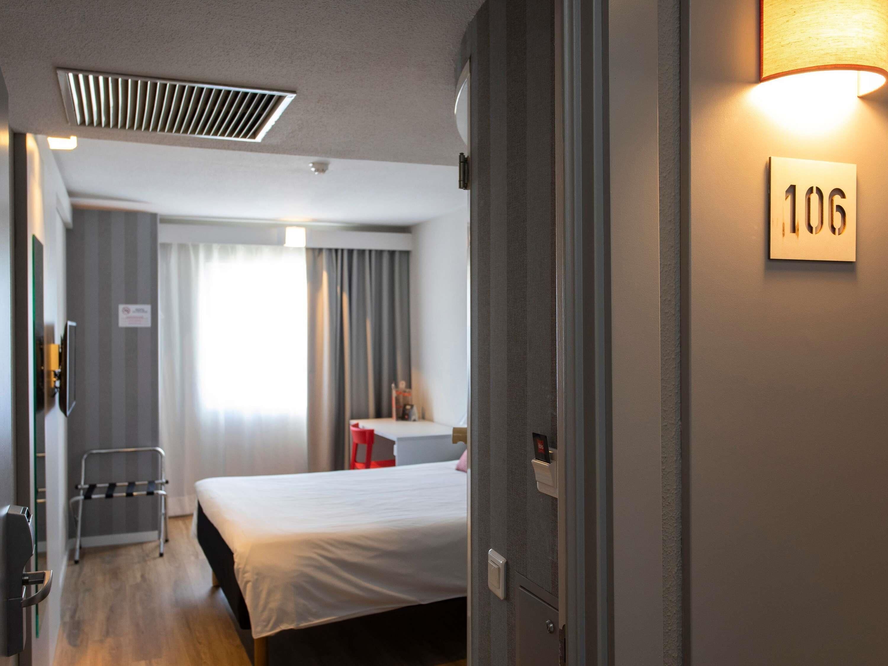 A hotel room with a bed, a desk, a chair, a window with curtains, and a wall-mounted number sign.