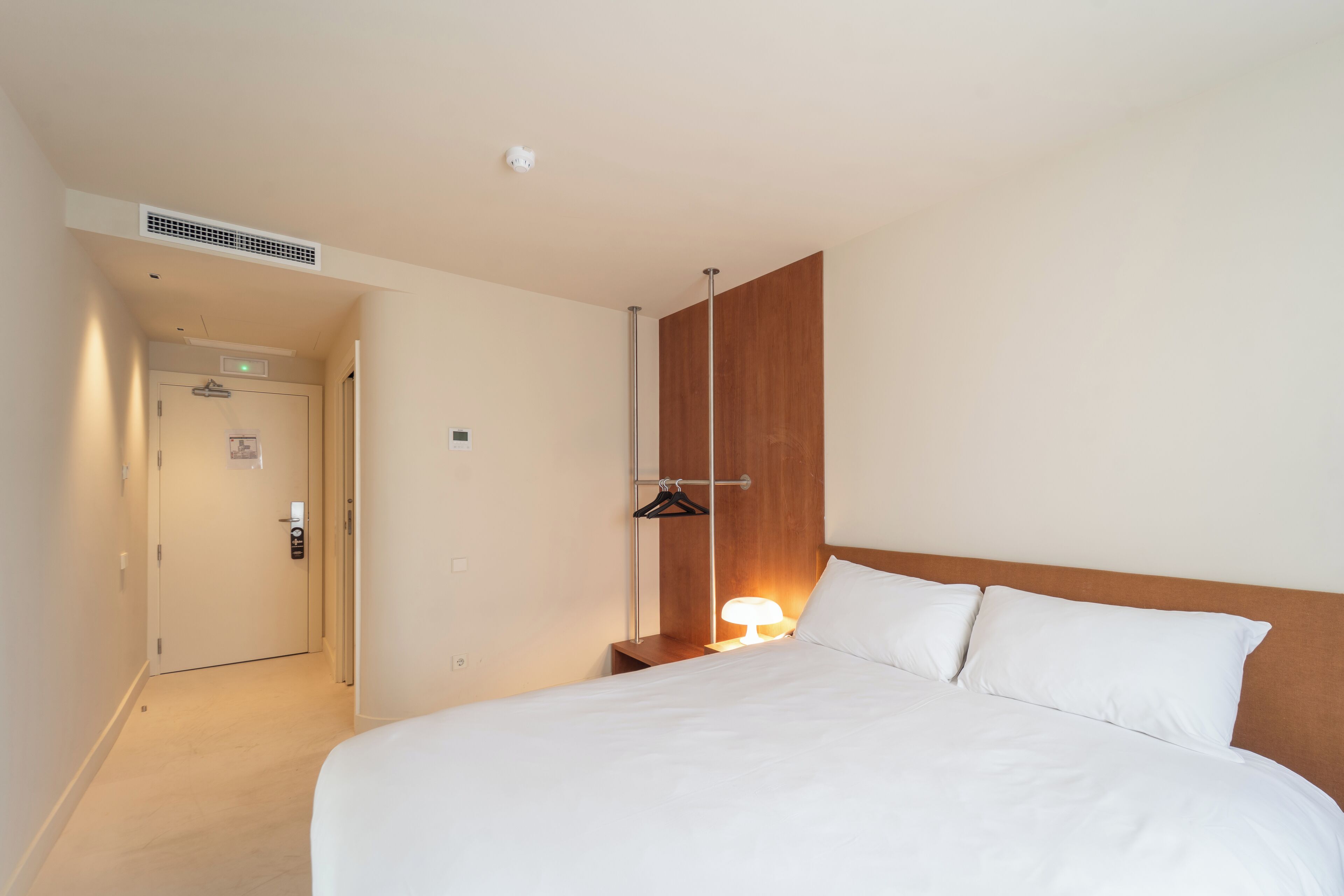 Habitación de hotel con una cama grande, una mesita de noche con lámpara, un cabecero de madera, una puerta con cerradura y un perchero en la pared.