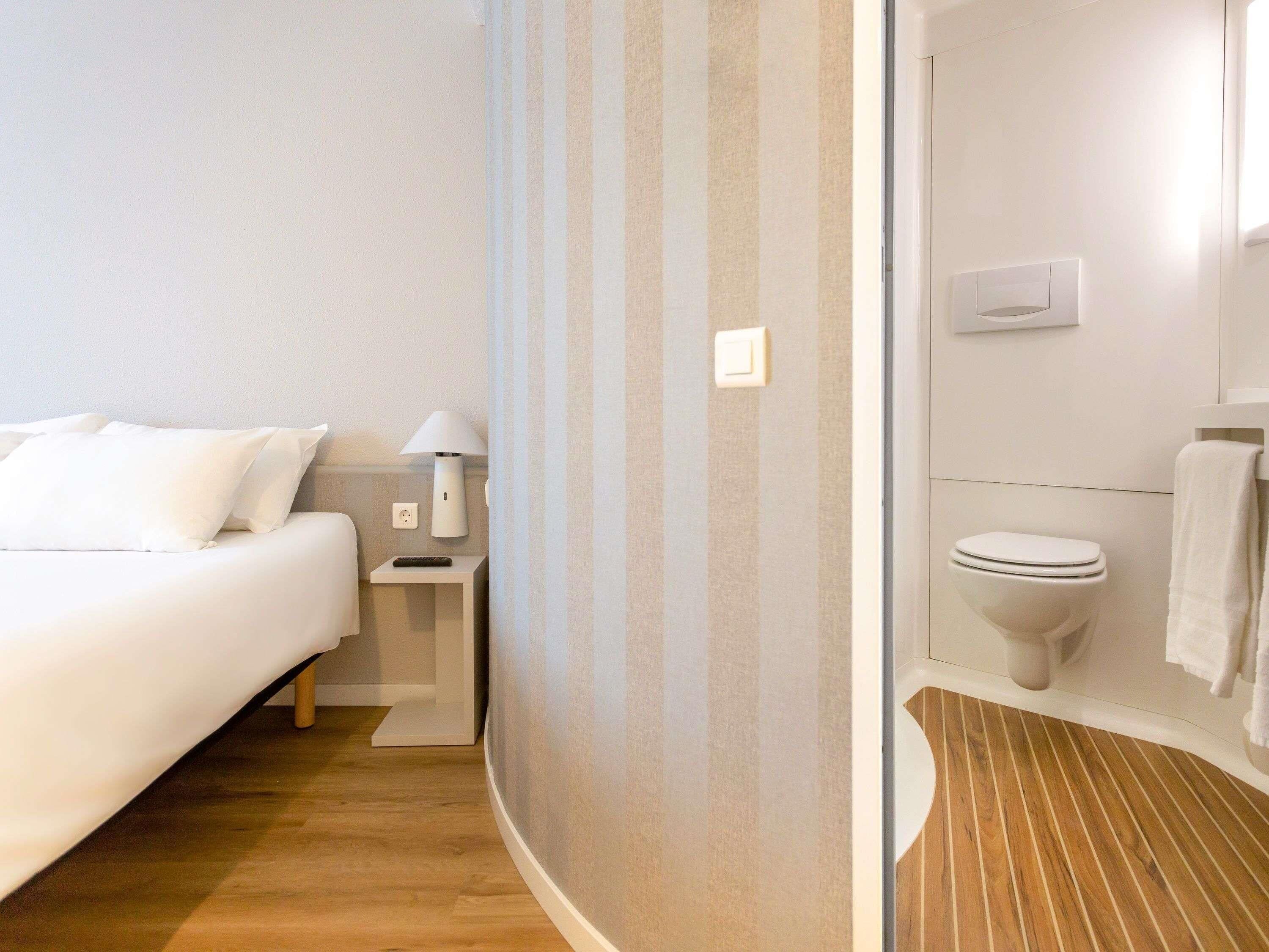 A hotel room with a bed, bedside table, lamp, and a bathroom with a toilet and towel rack.